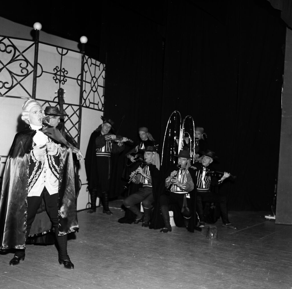 Svartvit bild på en teaterscen, till vänster på bilden står en man i peruk och sjunger, till höger flera andra skådespelare som tittar på.