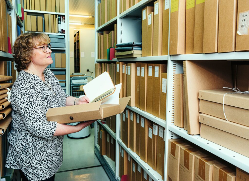 Kvinna till vänster på bilden håller i arkivmaterial, står framför arkivhyllor.